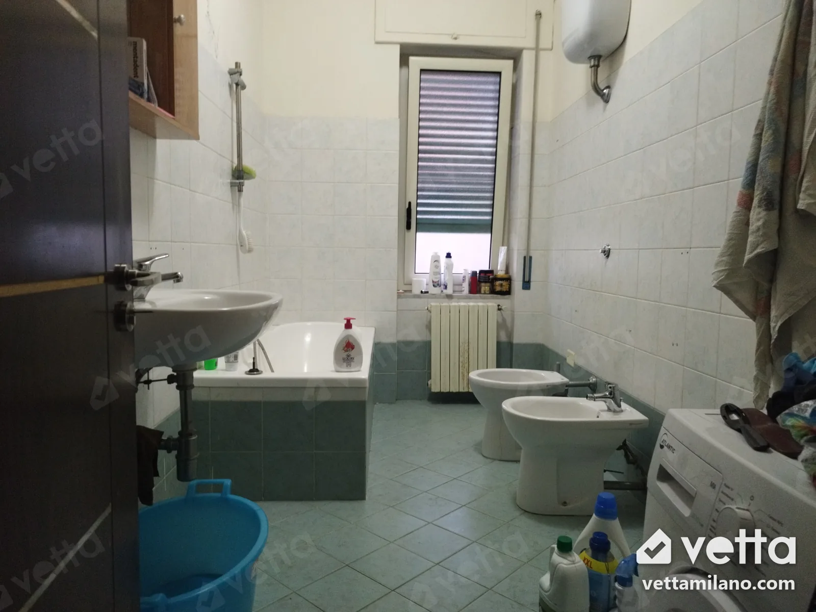 Foto di un bagno di casa com'è oggi: vasca lungo la parete, lavabo vicino alla porta, finestra in fondo, piastrelle chiare con lo zoccolo verde acqua.